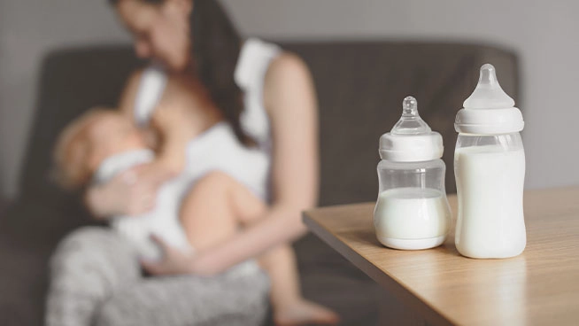 Two bottles in focus with mother in background breast feeding baby.