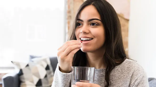 Una mujer sonriente con el pelo largo y oscuro sostiene un vaso de agua y está a punto de tomar una pastilla