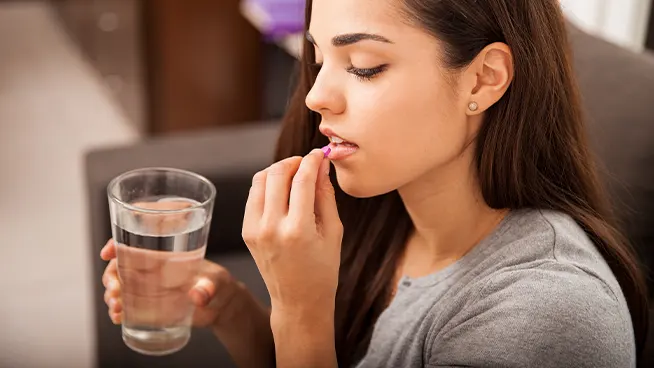 Una mujer con cabello largo y oscuro sostiene un vaso de agua y está a punto de tomar una pastilla