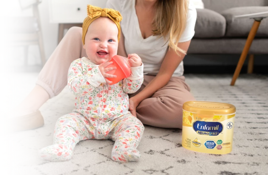 Baby smiling and holding bottle while sitting next to Mom on the floor