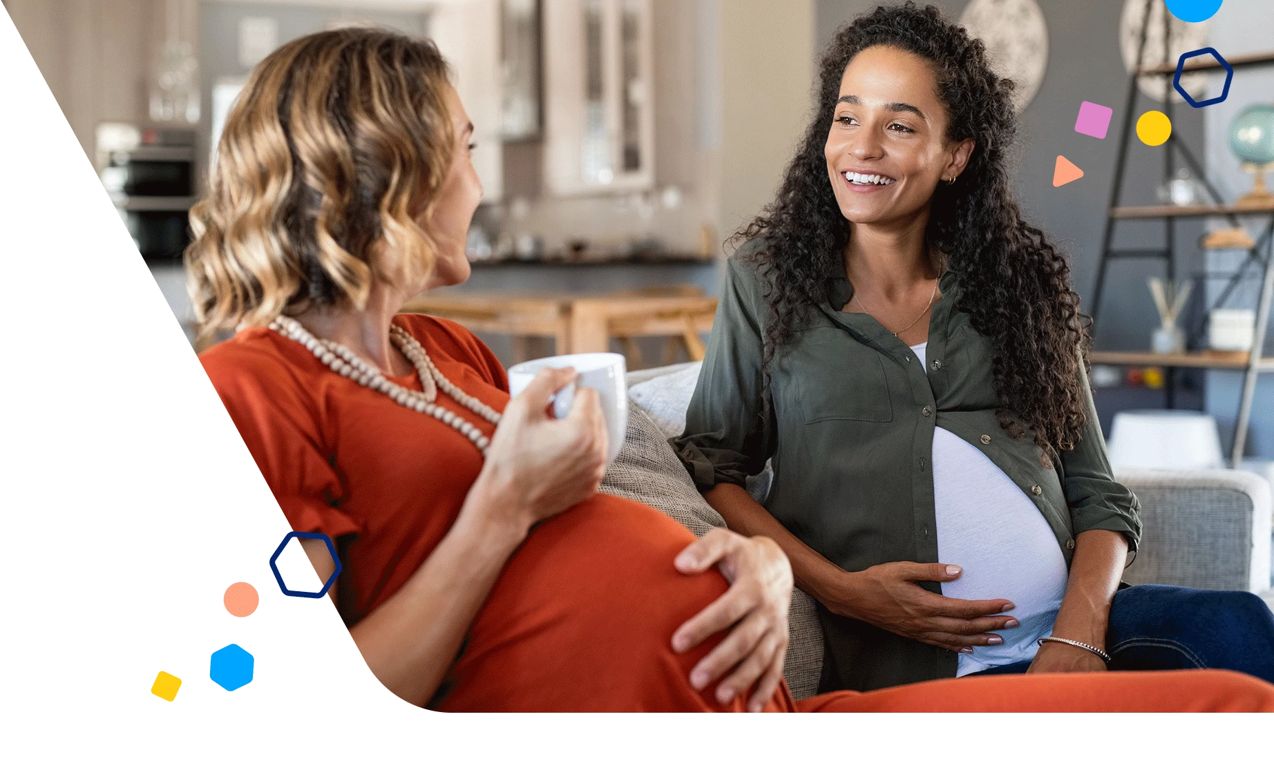 Two pregnant women sitting on the couch talking with one holding a cup of coffee