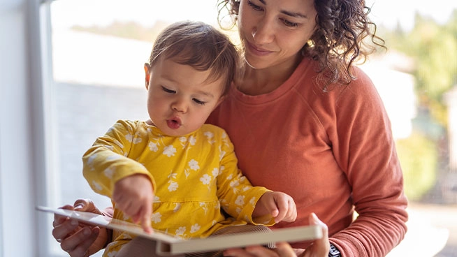 Mother with inquisitive toddler in lap looking through picture book.