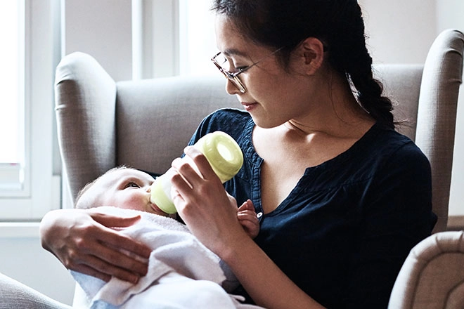 Smiling mother bottle feeding baby in chair.